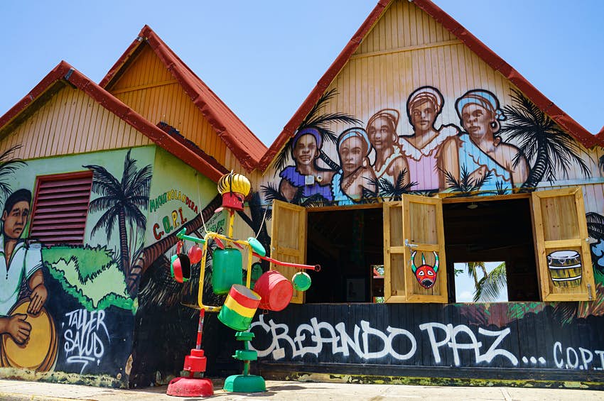 Un par de edificios están cubiertos con imágenes de mujeres con pañuelos en la cabeza y un hombre tocando un tambor;  frente al edificio hay una colorida estructura de ollas y sartenes;  Puerto Rico Sustentable 